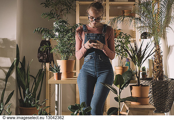 Woman using smart phone while leaning on table by potted plants at home