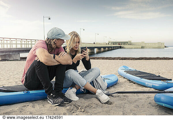 Woman using smart phone while male friend sitting by on paddleboard