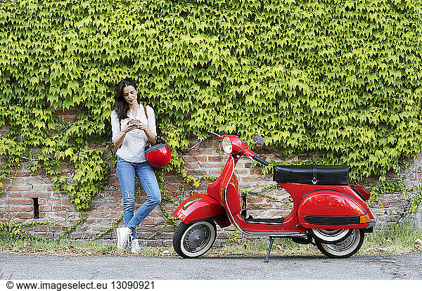 Woman using smart phone while standing by motor scooter against ivy wall