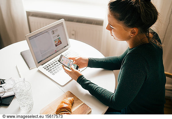 Woman using smart phone while surfing food recipe on laptop at home