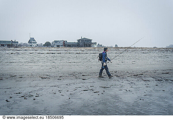 Woman walking across beach in snow with fly fishing equipment