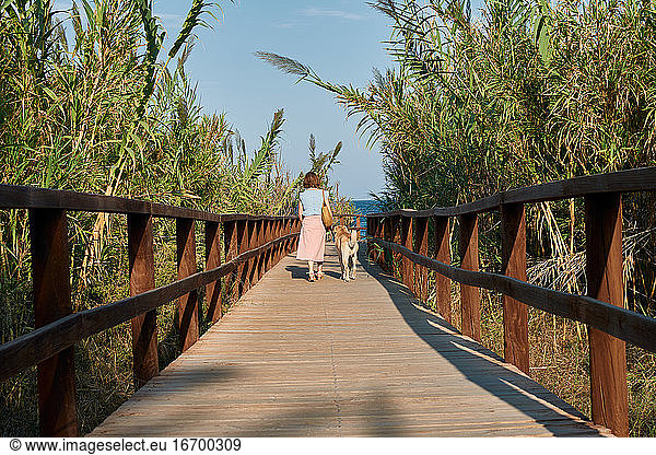 Woman walks with her dog on a wooden catwalk towards the sea
