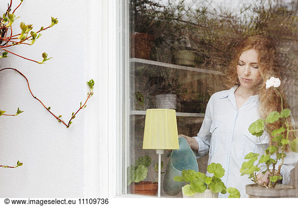 Woman watering potted plant at home seen from glass window