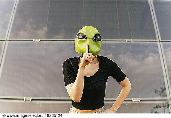 Woman wearing alien mask gesturing in front of glass building