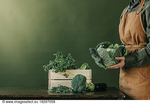Woman wearing apron holding cabbage in front of green wall