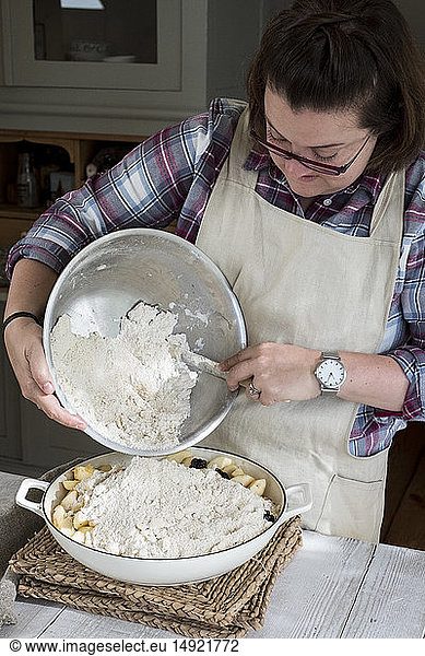 Woman wearing apron standing in a kitchen  pouring fresh crumble mixture on to a pie dish filled with fruit.