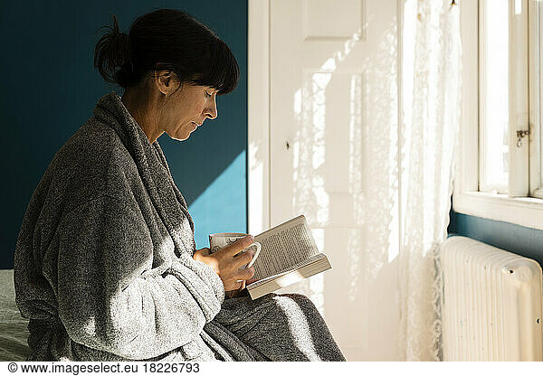 Woman wearing bathrobe holding cup while reading book in bedroom at home