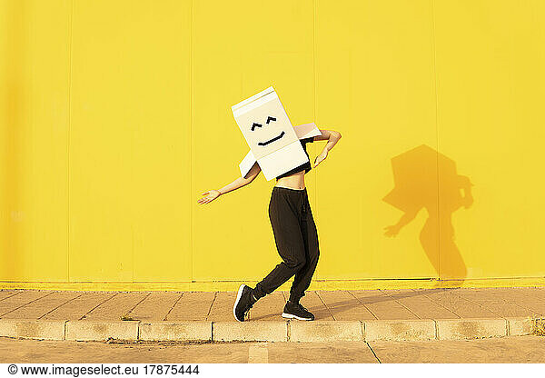 Woman wearing box with smiley face enjoying dance in front of of yellow wall on footpath