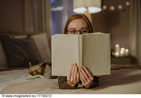 Woman wearing eyeglasses covering face with book on bed at home