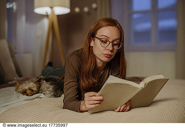 Woman wearing eyeglasses reading book lying by Yorkshire Terrier on bed at home