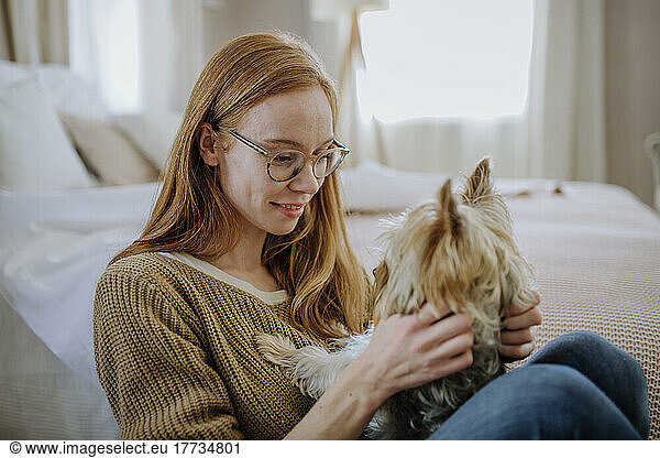 Woman wearing eyeglasses sitting with pet dog by bed at home