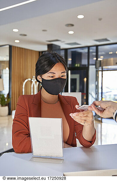 Woman wearing face mask taking cardkey from receptionist at hotel reception