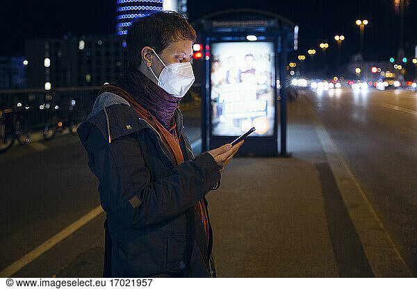 Woman wearing face mask using mobile phone while standing at bus stop in city