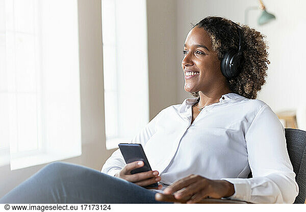 Woman wearing headphones looking away while sitting with mobile phone on armchair at home