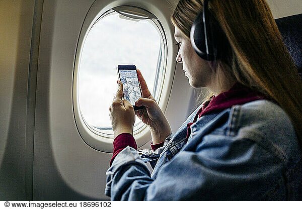 Woman wearing headphones photographing with mobile phone through airplane window