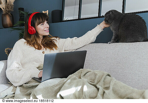 Woman wearing headphones stroking cat sitting on sofa at home