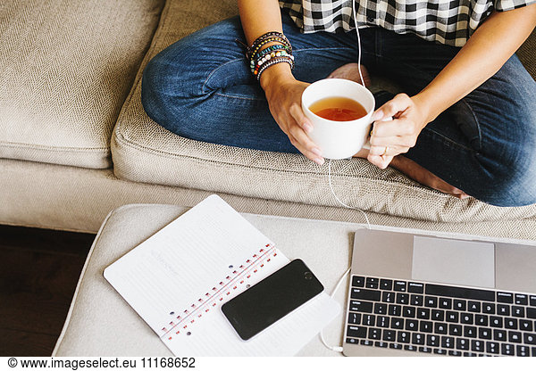 Woman wearing jeans sitting on a sofa in front of a laptop computer  holding a mug.