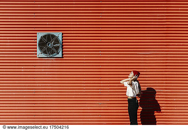 Woman wearing knit hat by red corrugated wall on sunny day