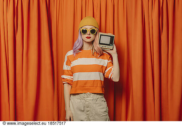 Woman wearing knit hat holding retro style television set in front of orange curtain