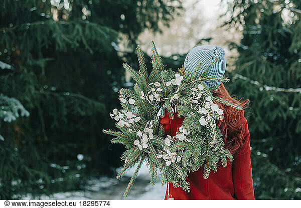 Woman wearing knit hat holding wreath in front of trees