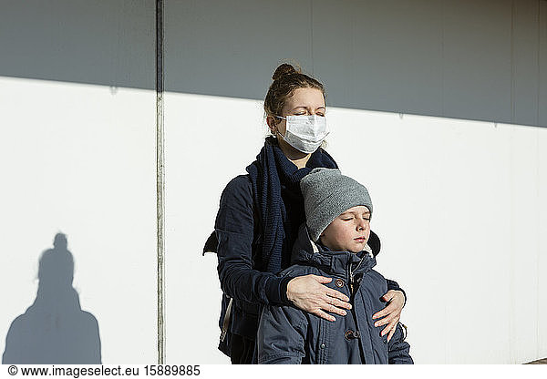 Woman wearing mask standing with son in front of a white wall  eyes closed