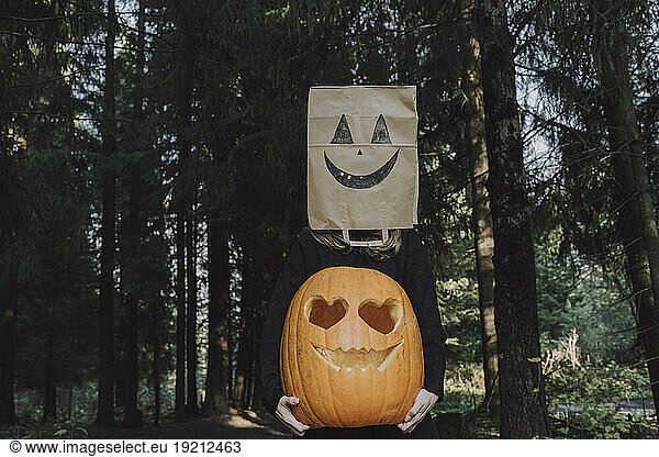 Woman wearing paper bag over face holding Jack O' Lantern in forest