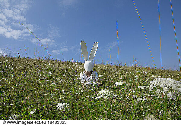 Woman wearing rabbit mask crouching in field under sky