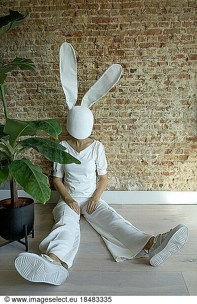 Woman wearing rabbit mask sitting near plant and brick wall