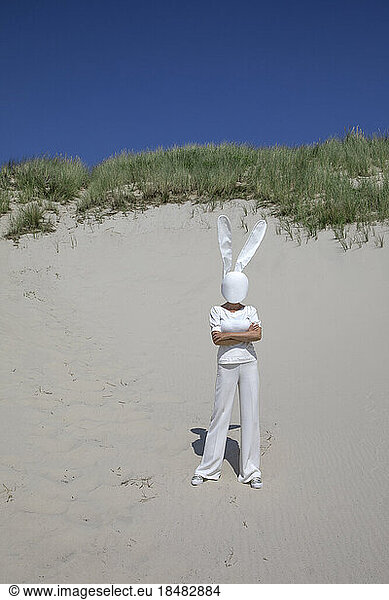 Woman wearing rabbit mask standing with arms crossed on sand dune
