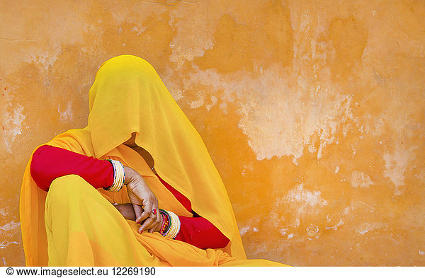 Woman wearing red and yellow sari and veil covering her head sitting on floor  leaning against orange wall.