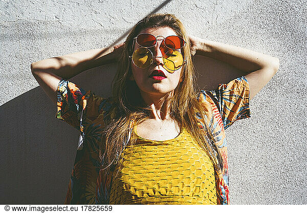 Woman wearing red and yellow sunglasses leaning with hands behind head on wall