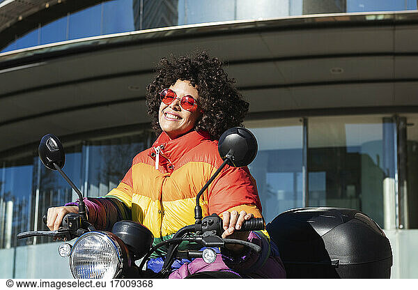 Woman wearing sunglasses and colorful jacket smiling while sitting on motorcycle