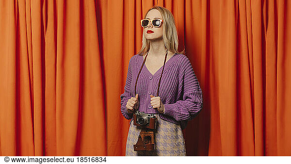 Woman wearing sunglasses holding vintage camera in front of orange curtain