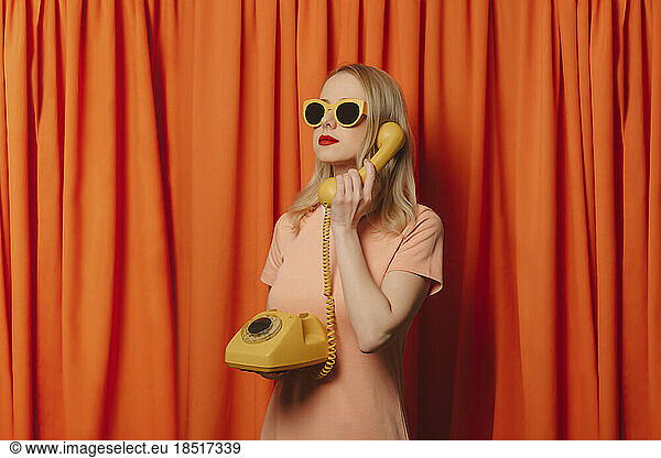 Woman wearing sunglasses talking over landline phone in front of orange curtains