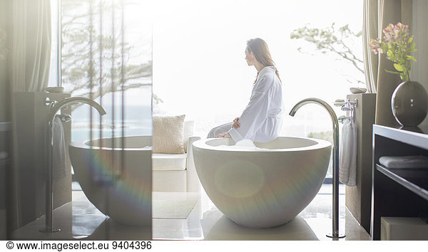Woman wearing white bathrobe  sitting on edge of bathtub and looking through window