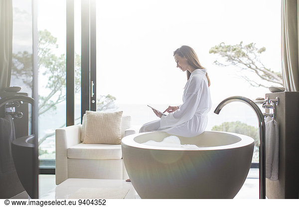 Woman wearing white bathrobe sitting on edge of modern bathtub and using digital tablet