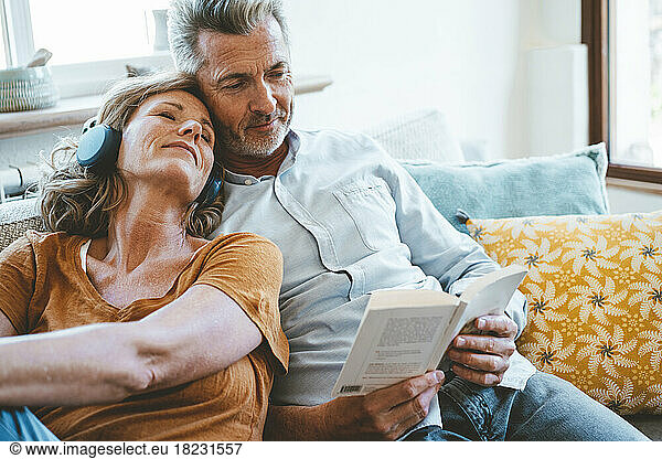 Woman wearing wireless headphones sitting by man reading book on sofa