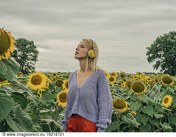 Woman wearing wireless headphones standing with eyes closed in sunflower field