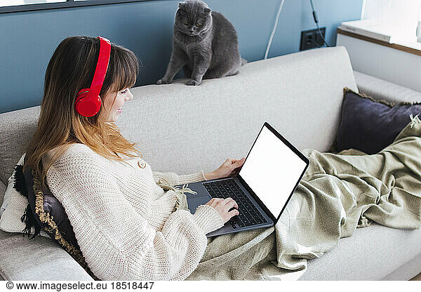 Woman wearing wireless headphones watching laptop on sofa at home
