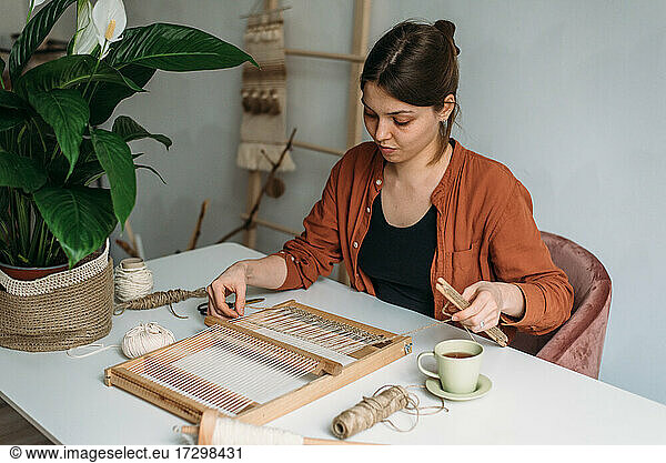 Woman weaves wool rug at white table with skeins and spathiphyllum