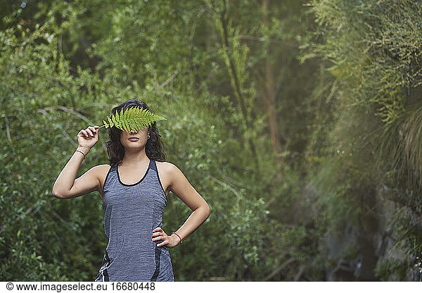 Woman with a fern on her face. She is in a river and covers half of her face with a fern leaf.