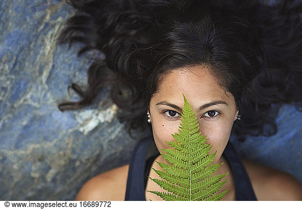Woman with a fern on her face. She's lying on a rock and covers half her face with a fern leaf.
