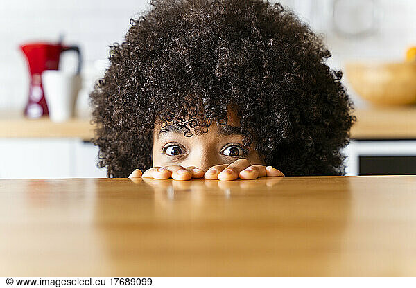 Woman with Afro hairstyle hiding behind kitchen island at home