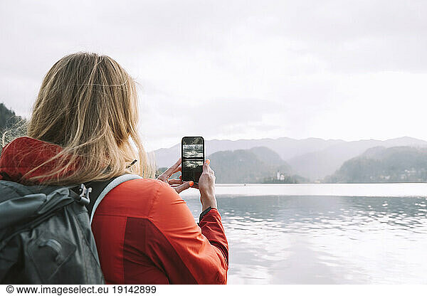 Woman with backpack clicking picture from smartphone near lake