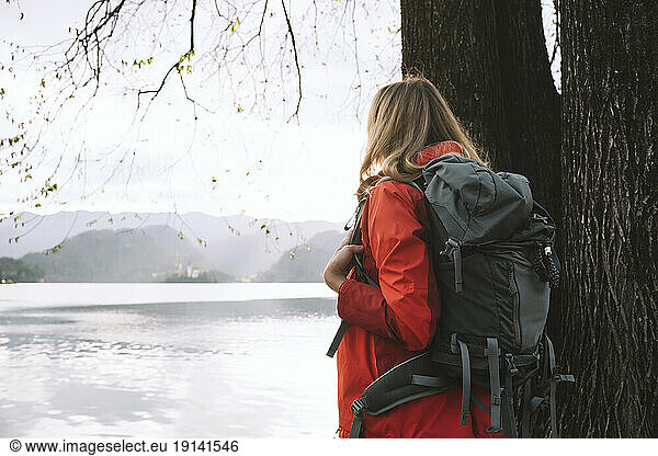 Woman with backpack standing next to tree near lake