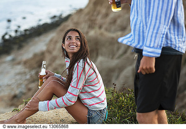 Woman with beer bottle looking at boyfriend while sitting on field