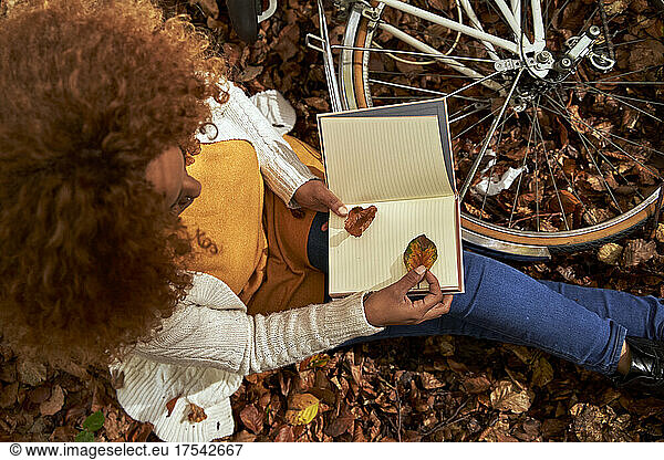 Woman with bicycle keeping leaf inside book sitting at autumn forest