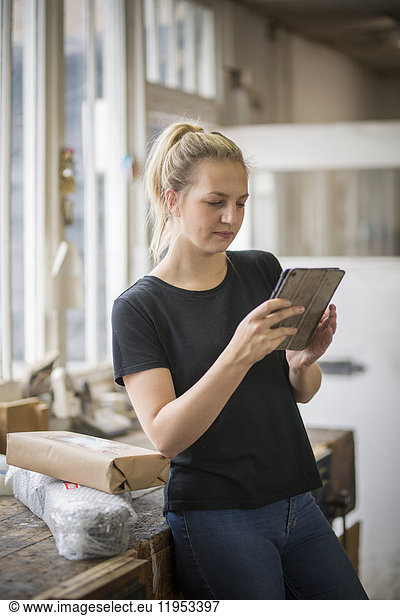Woman with blond hair standing in a workshop using a digital tablet  wrapped parcels on a table.
