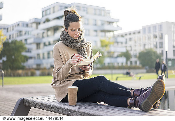 Woman with coffee to go sitting on bench in autumn reading a book