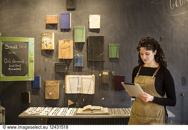 Woman with curly brown hair wearing apron standing in her pottery shop  looking at digital tablet  display of books and jewellery.